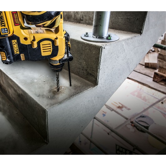 A yellow DEWALT rotary hammer drill is being used to drill into a concrete staircase step, with construction materials visible in the background.