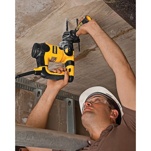 A person is using a DEWALT hammer drill to drill into a concrete ceiling. The person is wearing a safety helmet and work clothes. The yellow and black corded power tool is being operated above their head.