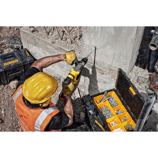 A construction worker wearing a yellow hard hat and an orange safety vest uses a DEWALT drill with a masonry bit to drill into a concrete wall outdoors. Next to him is an open DEWALT toolkit containing various drill bits and fasteners.