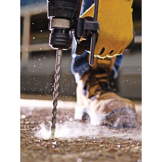 Close-up of a person wearing yellow gloves and brown work boots using a DEWALT drill bit to drill into a concrete surface, with dust and debris visible.