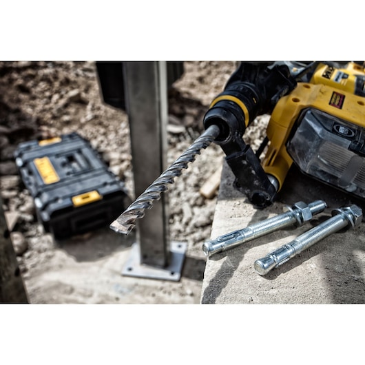 Close-up of a DEWALT power drill with a spiral drill bit, resting on concrete alongside three large metal anchor bolts, with a black DEWALT toolbox in the background at a construction site.