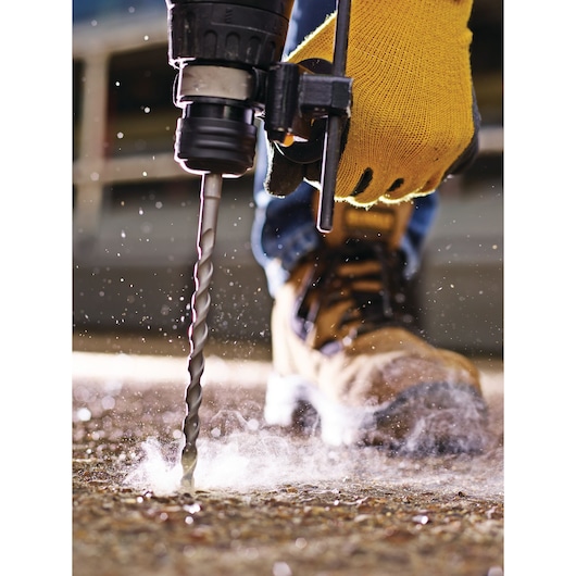 Close-up of a person wearing a yellow glove using a power drill with a twist drill bit on a concrete surface, with dust being generated. The person is also wearing work boots.