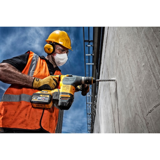 A construction worker wearing a yellow hard hat, orange safety vest, and gloves is using a DEWALT cordless rotary hammer drill with a DT9517 drill bit to drill into a concrete wall outdoors.