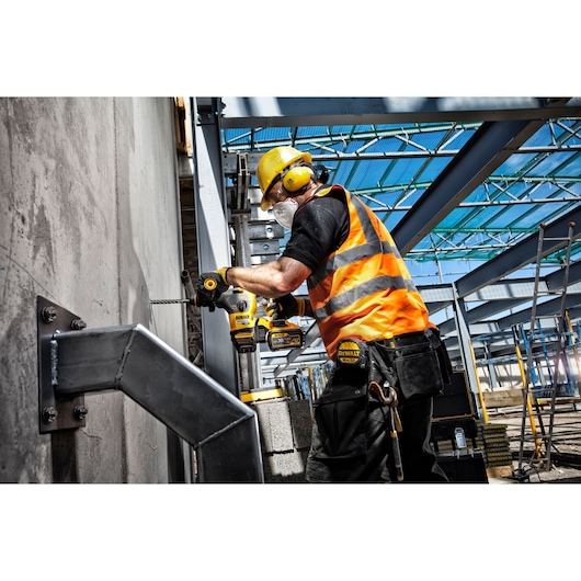 A construction worker wearing a yellow hard hat, ear protection, safety goggles, an orange reflective vest, and a face mask is using a DEWALT power drill to drill into a concrete wall at an industrial building site.