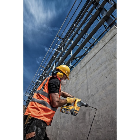 A construction worker wearing an orange safety vest, yellow helmet, protective mask, and ear protection is using a yellow DEWALT rotary hammer drill to drill into a concrete wall outdoors under a blue sky.