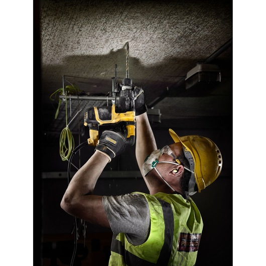 A construction worker wearing a yellow hard hat, safety vest, gloves, and face mask is using a DEWALT power drill with a long bit to drill into a ceiling. The worker is standing indoors and appears to be performing overhead work, emphasizing safety and heavy-duty equipment.