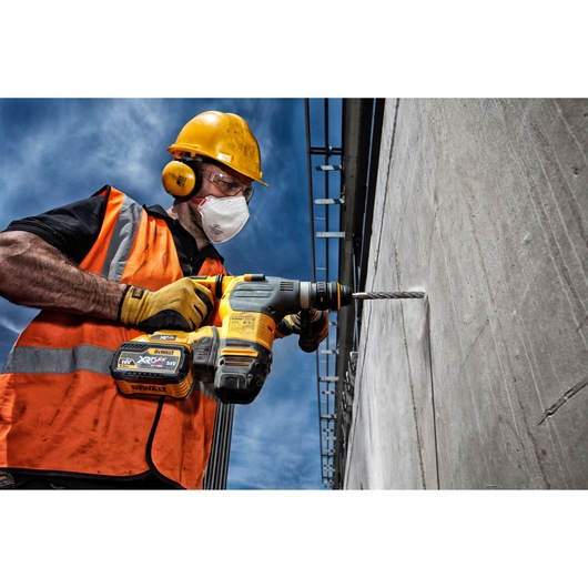 A construction worker wearing an orange safety vest, yellow helmet, and gloves is using a DEWALT cordless rotary hammer drill to drill into a concrete wall outdoors under a blue sky.