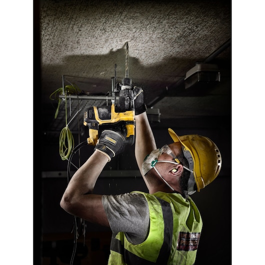 A construction worker wearing a safety helmet, gloves, protective vest, and face mask is using a yellow DEWALT rotary hammer