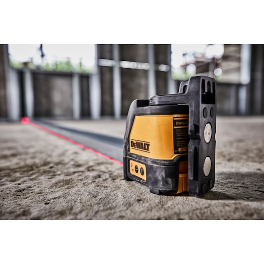 A DEWALT DW088K self-leveling cross line laser tool is shown projecting a red laser line on the floor inside an unfinished building with concrete walls.