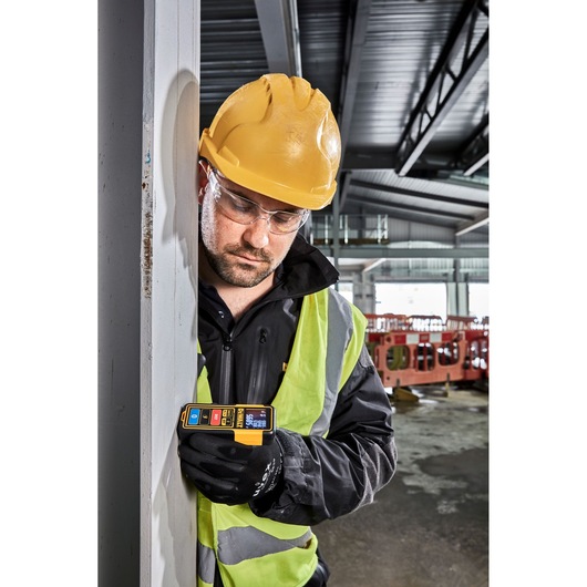 A construction worker wearing a yellow hard hat and reflective vest holds a DEWALT DW099S laser distance measurer against a