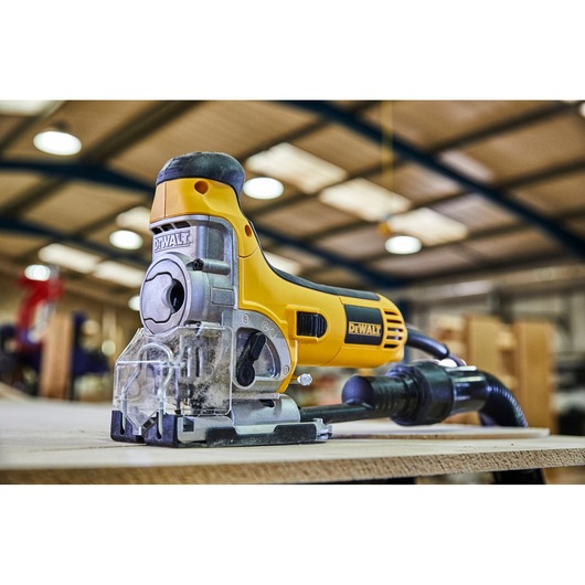 DEWALT electric jigsaw power tool in yellow and black, positioned on a wooden surface in a workshop setting. The tool is plugged in and appears ready for use.