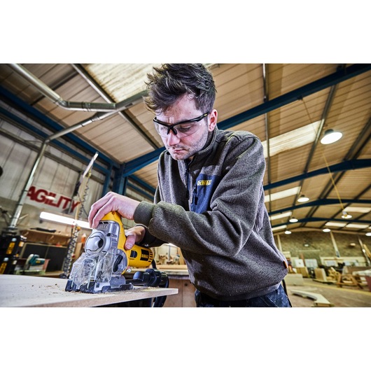 A person using a DEWALT DW333KT jigsaw to cut wood in a workshop. The individual’s face is blurred for privacy, and the workspace has high ceilings and industrial lighting.