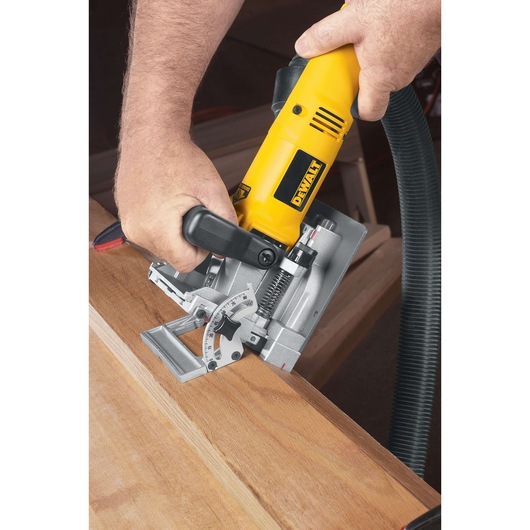 A person is using a DEWALT biscuit joiner power tool to cut a slot into a wooden board. The tool is yellow and black, and there is a vacuum hose attached for dust extraction.