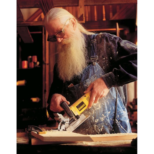 A person wearing denim overalls uses a DEWALT DW682K-QS biscuit joiner to cut slots into a piece of wood in a workshop setting. Wood shavings are visible, and the joiner's yellow body is prominent.