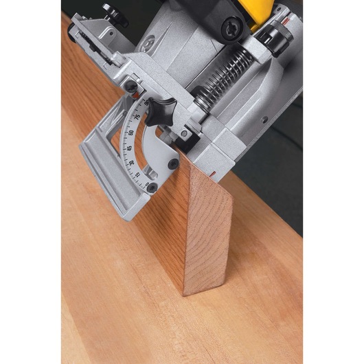 Close-up of a DEWALT electric biscuit joiner tool being used to cut a slot in a piece of wood at an angle. The tool shows a precise angle adjustment gauge and is positioned against a wooden surface.