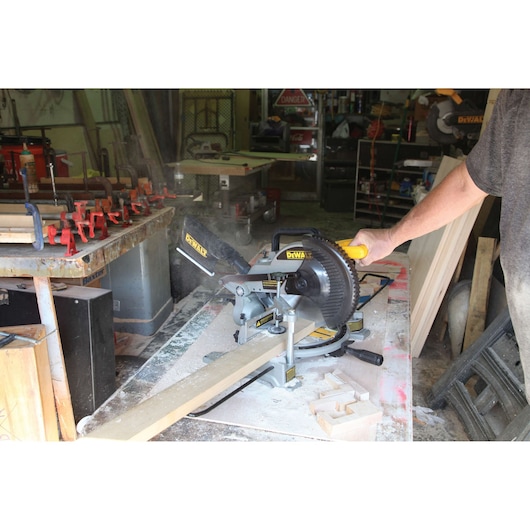 A person operating a DEWALT miter saw (SKU: DW714) to cut a wooden board in a workshop. Wood dust and clamps are visible on the worktable, with various tools and materials in the background.