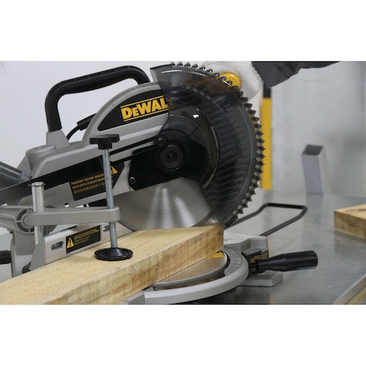 A DEWALT miter saw is being used to cut a piece of wood on a metal workbench. A person wearing a glove is operating the saw.