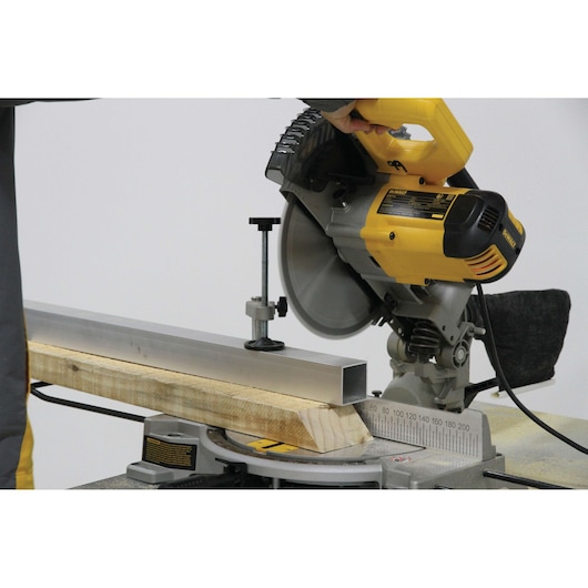 A yellow DEWALT miter saw being used to cut a piece of metal and a piece of wood, held securely in place on the saw’s base.