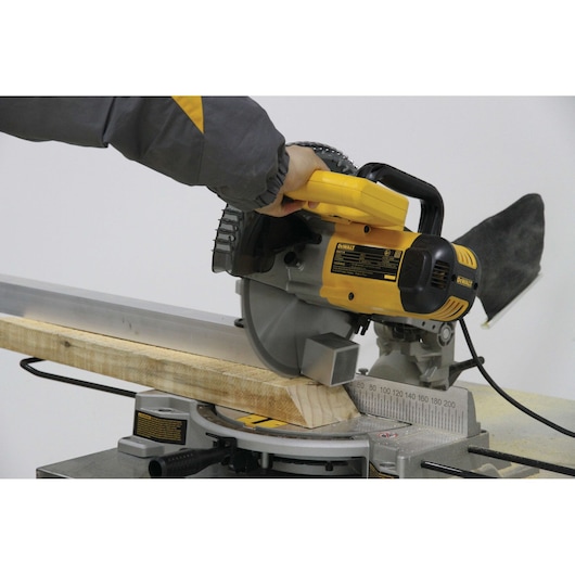 A DEWALT DW714 miter saw is being used to cut a piece of wood at an angle. The saw is yellow and black, and a person wearing a jacket is operating it.