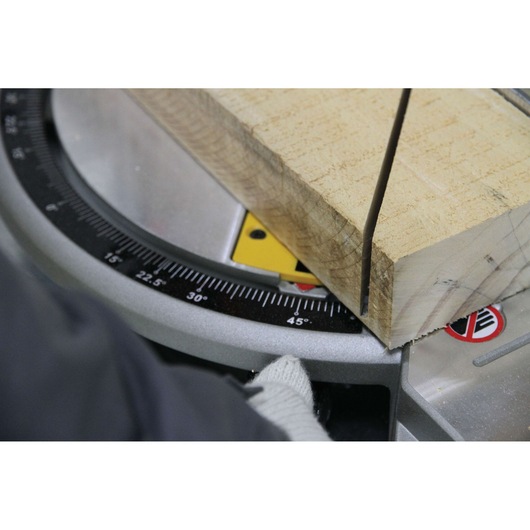 Close-up of a DEWALT miter saw cutting a piece of wood at a 45-degree angle. A gloved hand is visible adjusting the saw. The saw's angle gauge and safety markings are in view.
