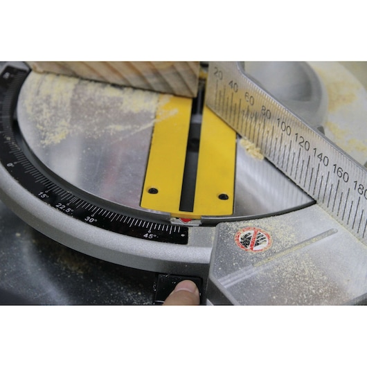 Close-up view of a DEWALT miter saw set at a 45-degree angle, with a piece of wood placed against the fence. Sawdust is visible on the surface and there is a warning sticker on the base.
