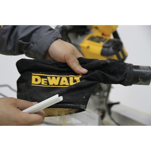 Close-up of a person holding a black DEWALT dust bag attached to a power tool, with another hand holding a white plastic piece.