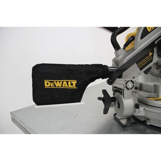 Close-up of a DEWALT miter saw with a black dust collection bag attached, positioned on a metal surface.