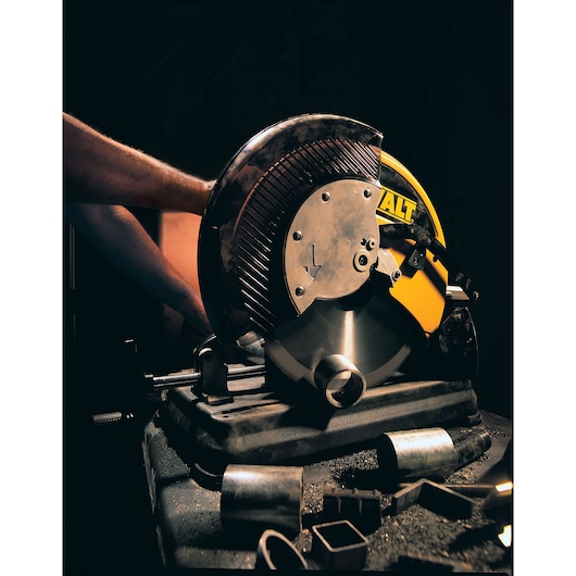 A close-up view of a DEWALT DW872 metal cutting saw in use, with a spinning blade cutting through a metal pipe, and various metal pieces and shavings on the work surface.