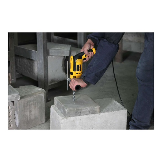 A person using a yellow DEWALT electric drill to drill a hole into a concrete block. The scene takes place in an industrial setting with additional concrete blocks in the background.