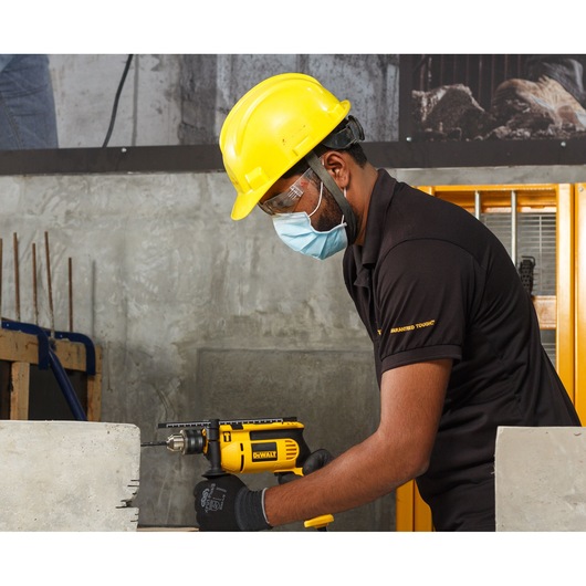 A worker wearing a yellow hard hat and black gloves uses a DEWALT DWD024 drill to work on a concrete block in an industrial setting. The worker's face is blurred for privacy.
