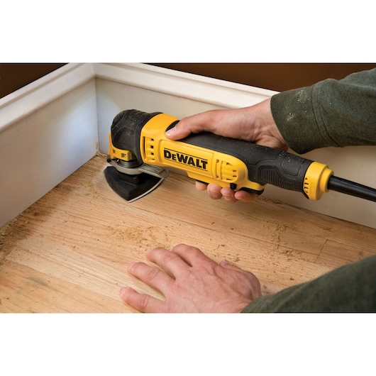 A person using a DEWALT DWE315KT oscillating multi-tool to sand wood flooring in a corner near a baseboard.