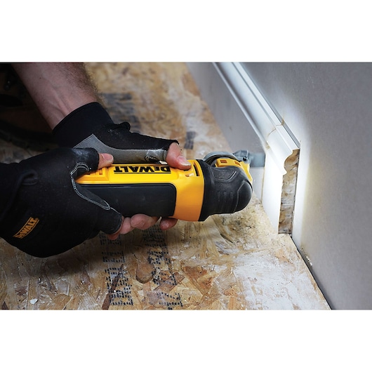 A person wearing gloves uses a DEWALT DWE315KT oscillating multi-tool to cut into a section of wood flooring and wall trim.