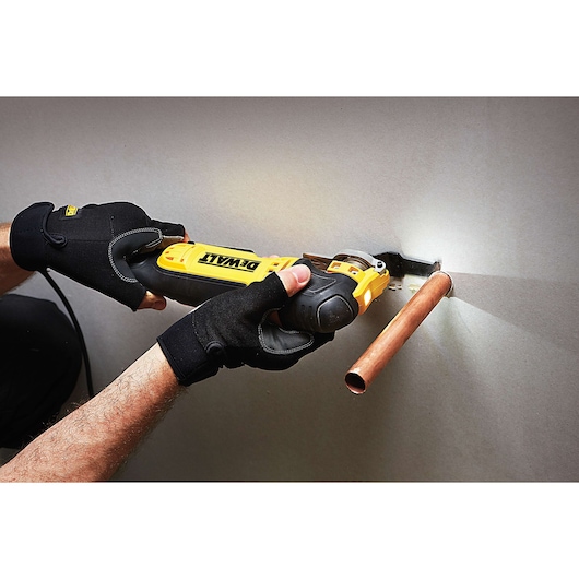 A person wearing black gloves uses a DEWALT multi-tool to cut drywall around a copper pipe.