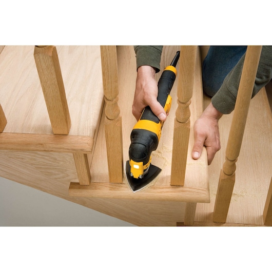 A person uses a DEWALT DWE315KT oscillating multi-tool to sand a wooden staircase, focusing on a difficult-to-reach area