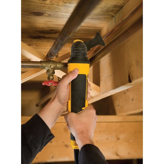 A person uses a DEWALT DWE315KT oscillating multi-tool to cut a pipe in a wooden basement or ceiling area.