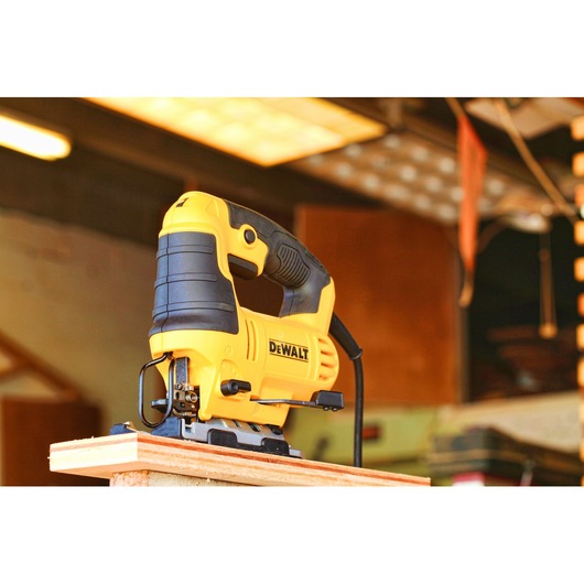 A yellow and black DEWALT electric jigsaw is positioned on the edge of a piece of wood in a workshop setting.