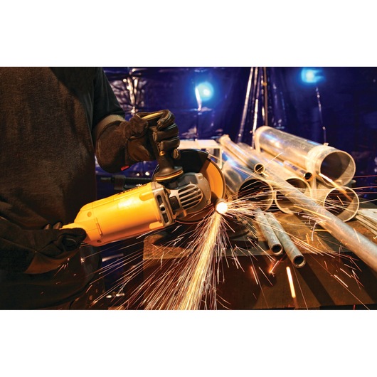 A person using a DEWALT angle grinder to cut metal pipes on a workbench, with bright sparks flying from the cutting area. Several metal pipes are stacked on the table in an industrial setting.