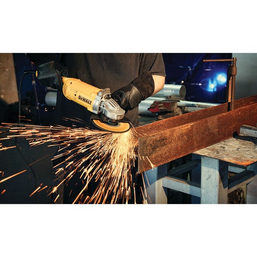A person using a yellow DEWALT angle grinder to cut or grind a metal beam, with sparks flying from the contact point. The person is wearing protective gloves and working in a workshop setting.