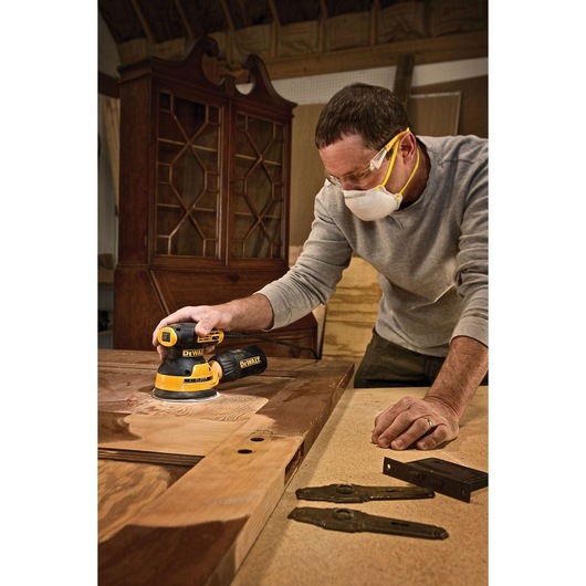 A person using a DEWALT DWE6423 random orbital sander to sand a wooden door in a woodworking shop. Some metal hardware pieces are positioned on the table nearby, and the person is wearing a protective mask.