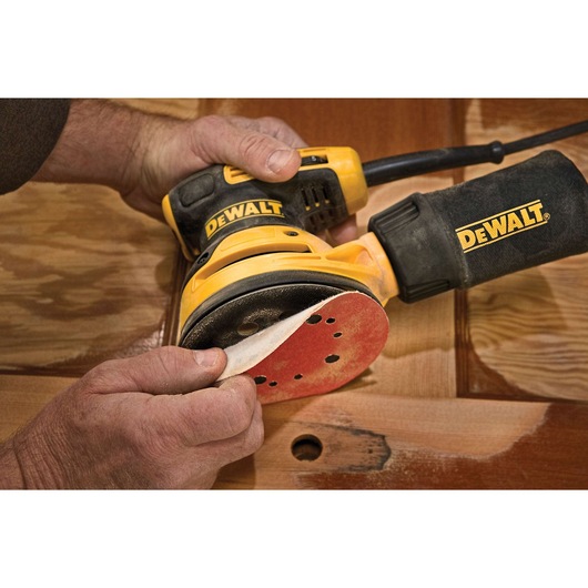 A person is changing the sandpaper pad on a DEWALT electric orbital sander, which is being used to work on a wooden surface.