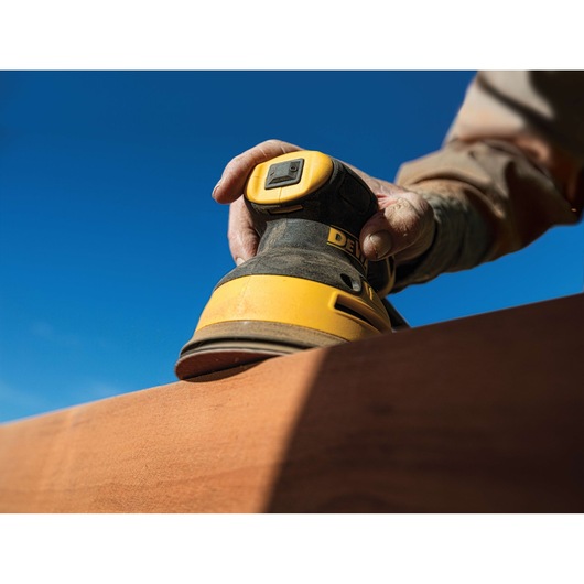 A close-up of a hand using a DEWALT DWE6423 yellow and black electric sander to smooth a wooden surface outdoors under a clear blue sky.