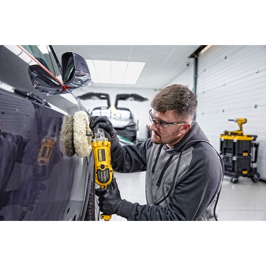 A person using a DEWALT DWP849X polisher to buff the side of a dark-colored car inside a garage. The person is wearing black gloves and a grey hoodie. The interior of the garage is bright, with tools and other equipment visible in the background.