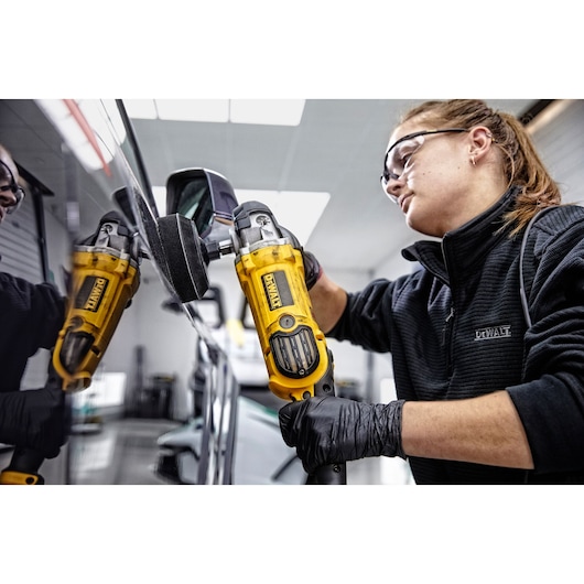 A person wearing black gloves and a DEWALT jacket is using a yellow DEWALT DWP849X polisher on the side of a car, with the reflection of the tool visible in the car's surface. The person's face is blurred.