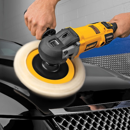 A DEWALT DWP849X rotary polisher in use, held by a person polishing the surface of a black car hood in a workshop setting.