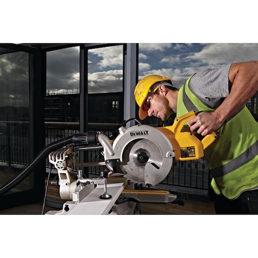 Worker uses a DEWALT DWS778 saw on a jobsite, wearing safety gear and operating the tool indoors near windows.