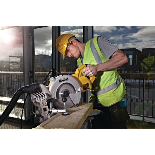 A person wearing a yellow hard hat and high-visibility vest using a DEWALT DWS778 miter saw to cut a wooden plank indoors
