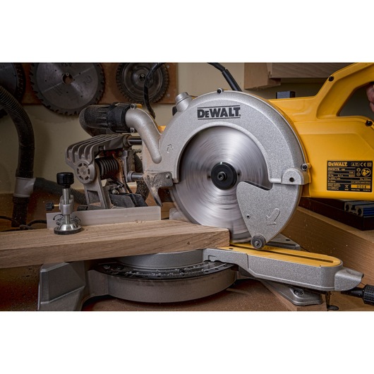 A DEWALT DWS778 sliding mitre saw cutting a piece of wood in a workshop, with the blade spinning and sawdust visible.