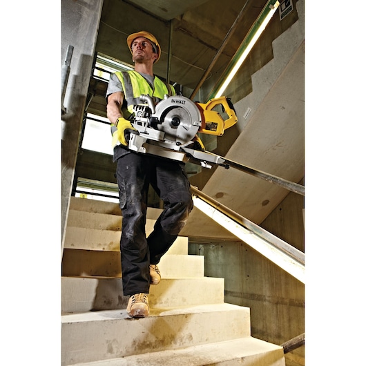 Worker in safety gear carries DEWALT DWS778 saw in a stairwell on a construction site.