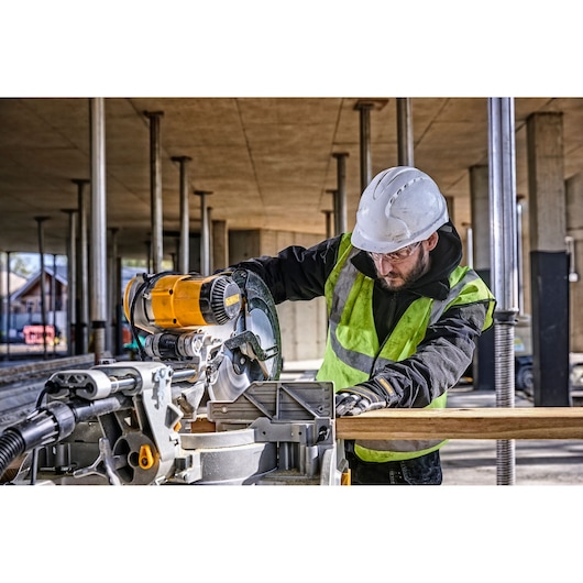 A person wearing a high-visibility vest and a hard hat is using a DEWALT DWS780 miter saw to cut a piece of wood at a construction site. The background shows structural columns and an unfinished building.