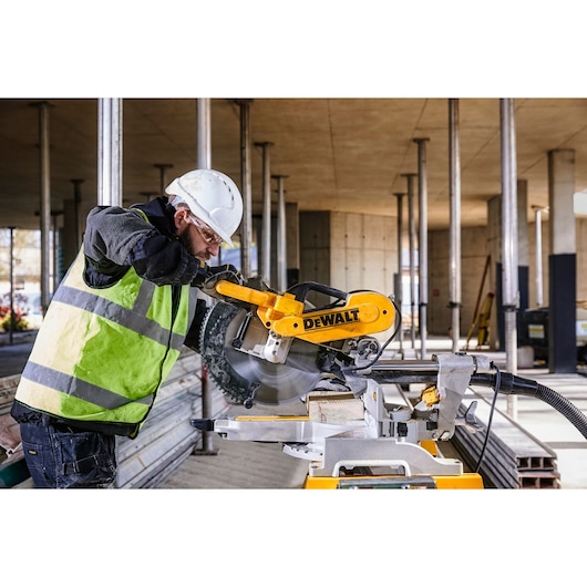 A construction worker wearing a helmet and high-visibility vest operates a DEWALT DWS780 miter saw on a building site. The saw is cutting a piece of wood, and there are metal columns and construction materials in the background.